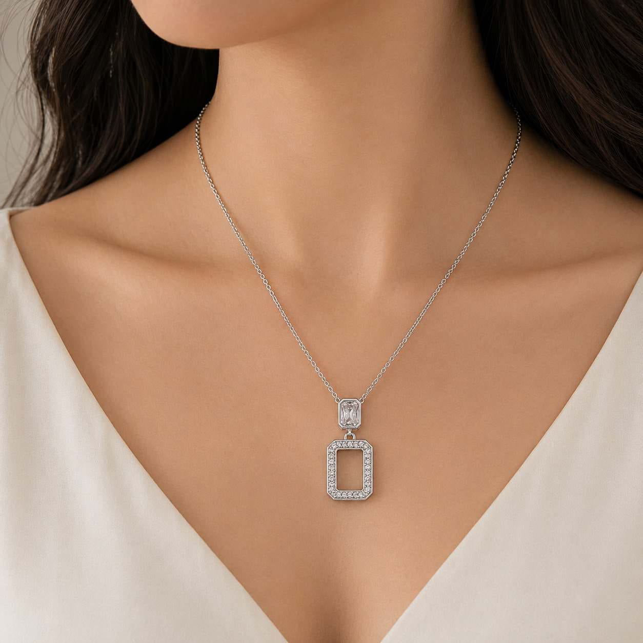 A close-up of a model wearing a delicate sterling silver chain necklace with a rectangular pendant with clear stones. 