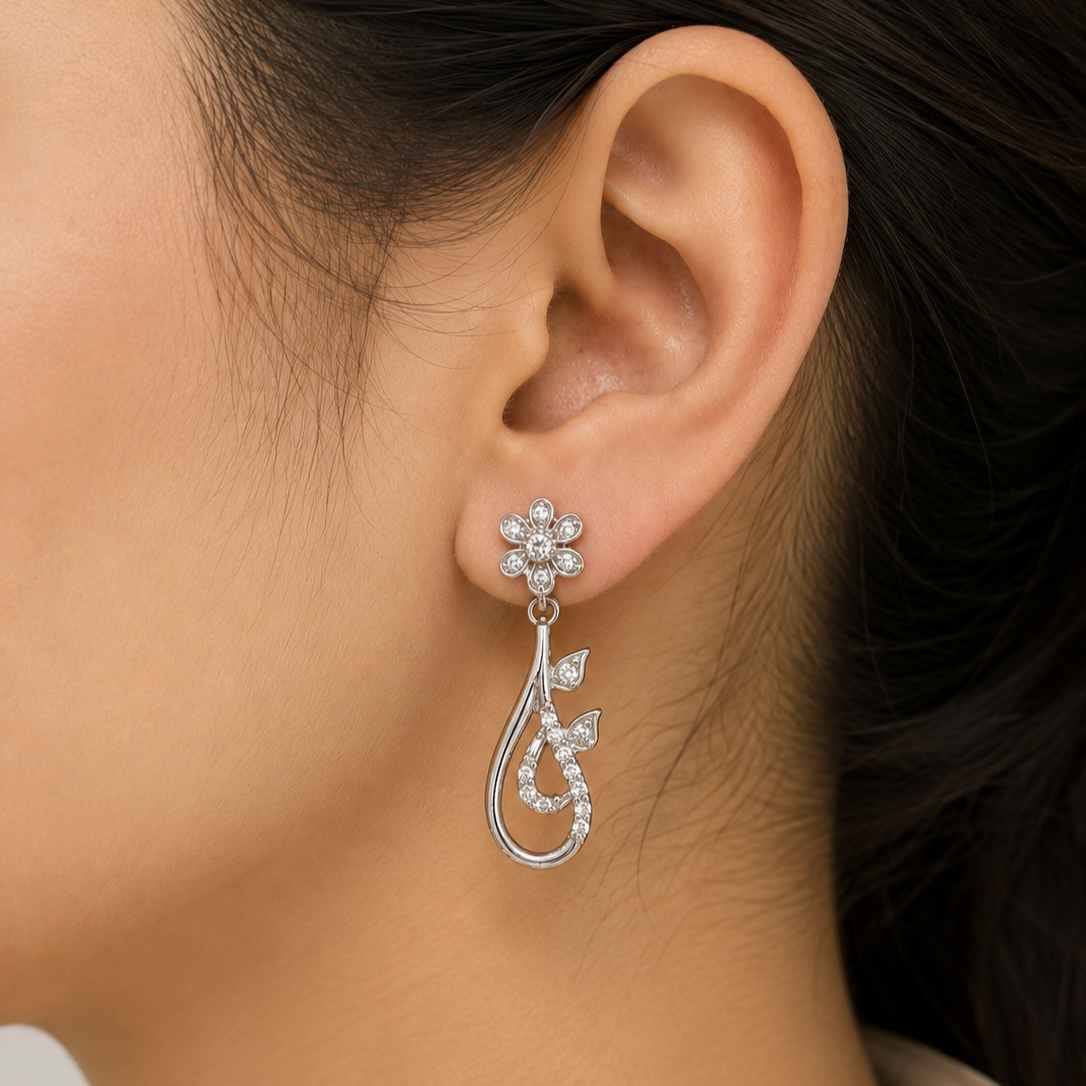 A close-up of a model's ear wearing a sterlinf silver dangle earring. The earring features a small flower-shaped stud set with clear stones, hanging from a curved, leaf-inspired drop pavé-set with clear stones.