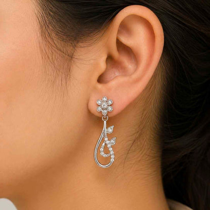 A close-up of a model's ear wearing a sterlinf silver dangle earring. The earring features a small flower-shaped stud set with clear stones, hanging from a curved, leaf-inspired drop pavé-set with clear stones.